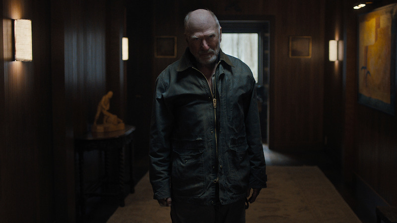 An older man with a bald head stands in a dimly lit, wood-paneled hallway with artwork on the walls and soft lighting, looking down slightly as light streams in from an open doorway behind him.
