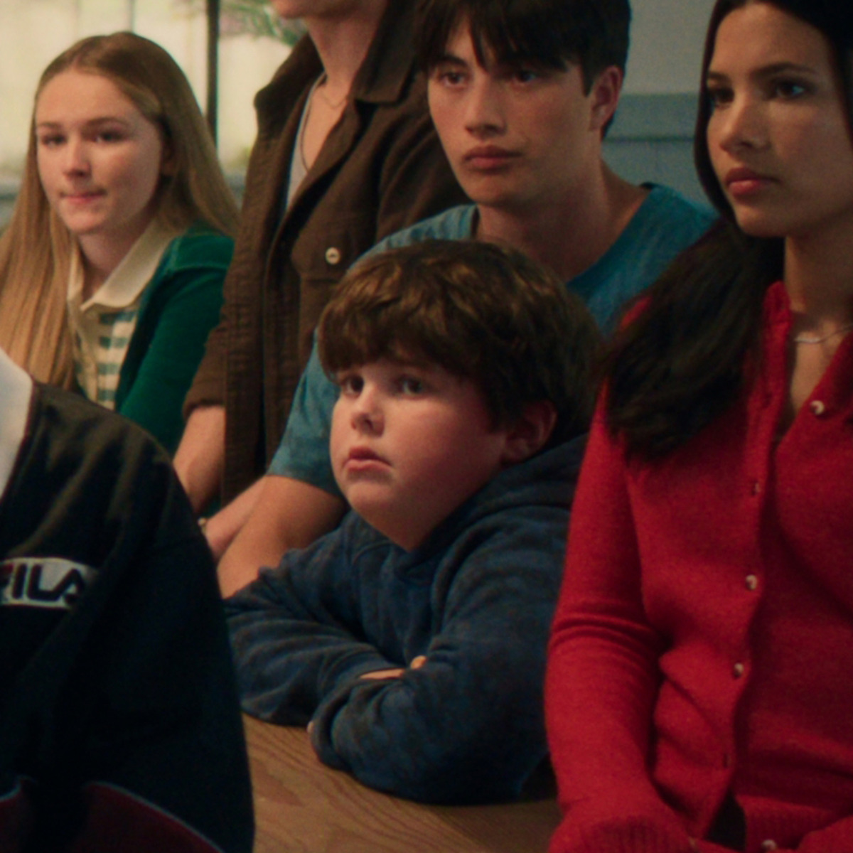 A group of teens and a child sit together at a table, listening attentively. The environment is casual with mixed expressions.