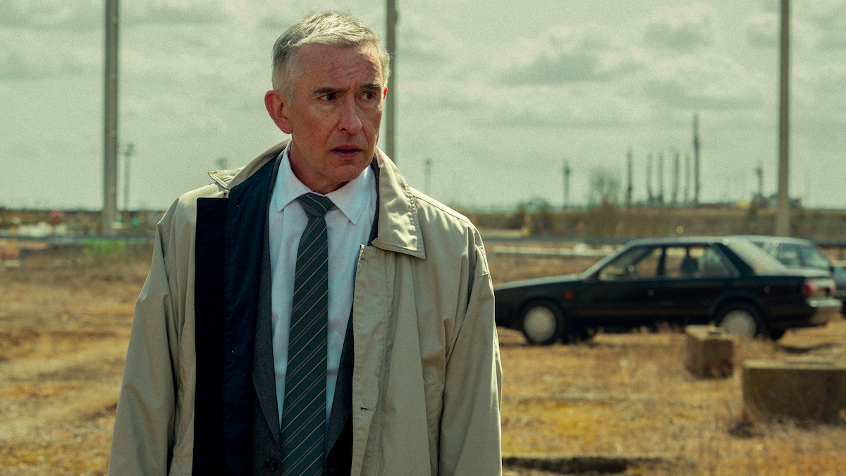 Middle-aged man in a trench coat and tie stands in an empty, industrial field with a dark car and refinery structures in the background, under a cloudy sky.
