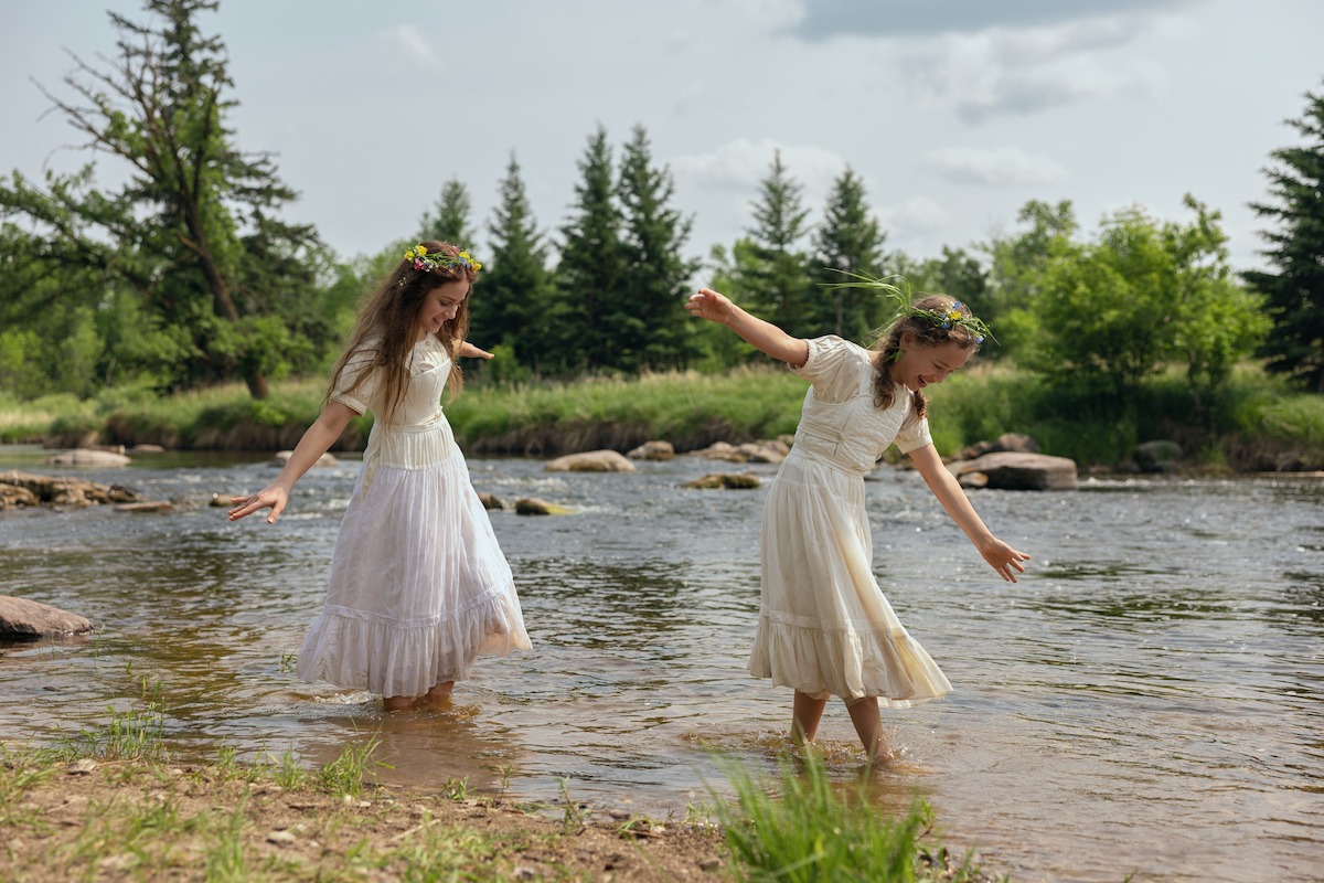 Two girls in white dresses with flower crowns wade and play in a shallow river surrounded by grass and trees on a bright, clear day.