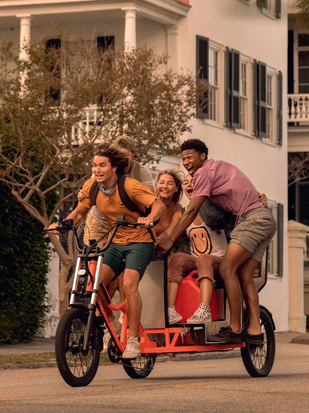 Four young friends ride a cargo bike joyfully on a suburban street in daylight, surrounded by greenery and classic white houses, capturing a moment of fun and carefree adventure.