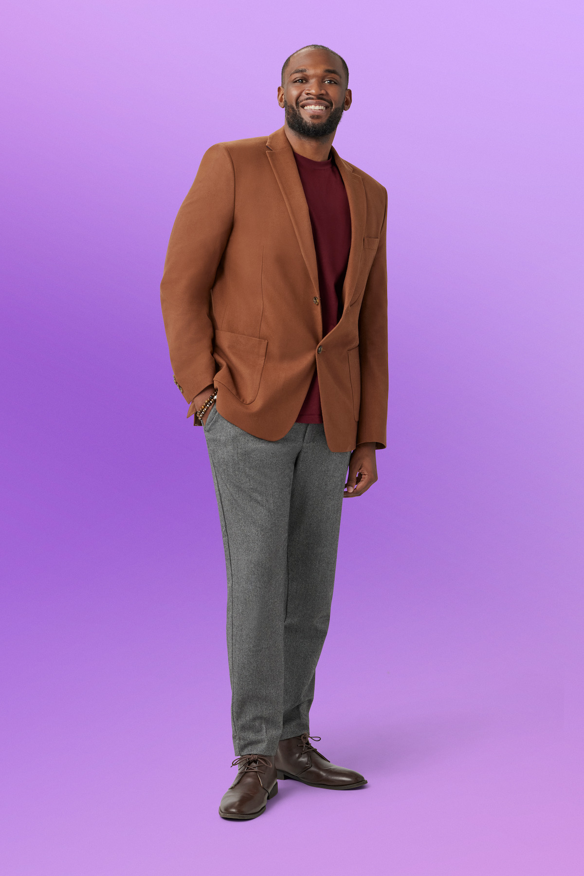 Kevan standing and smiling, wearing a brown blazer, maroon shirt, gray pants, and brown shoes, in front of a plain light purple studio background.