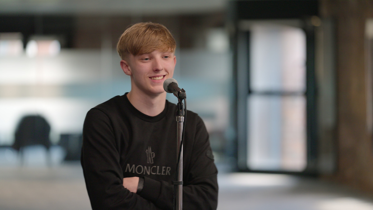 Young man in a Moncler sweatshirt stands smiling at a microphone in a modern, softly lit indoor space with glass doors and blurred furniture, suggesting a relaxed and casual atmosphere.