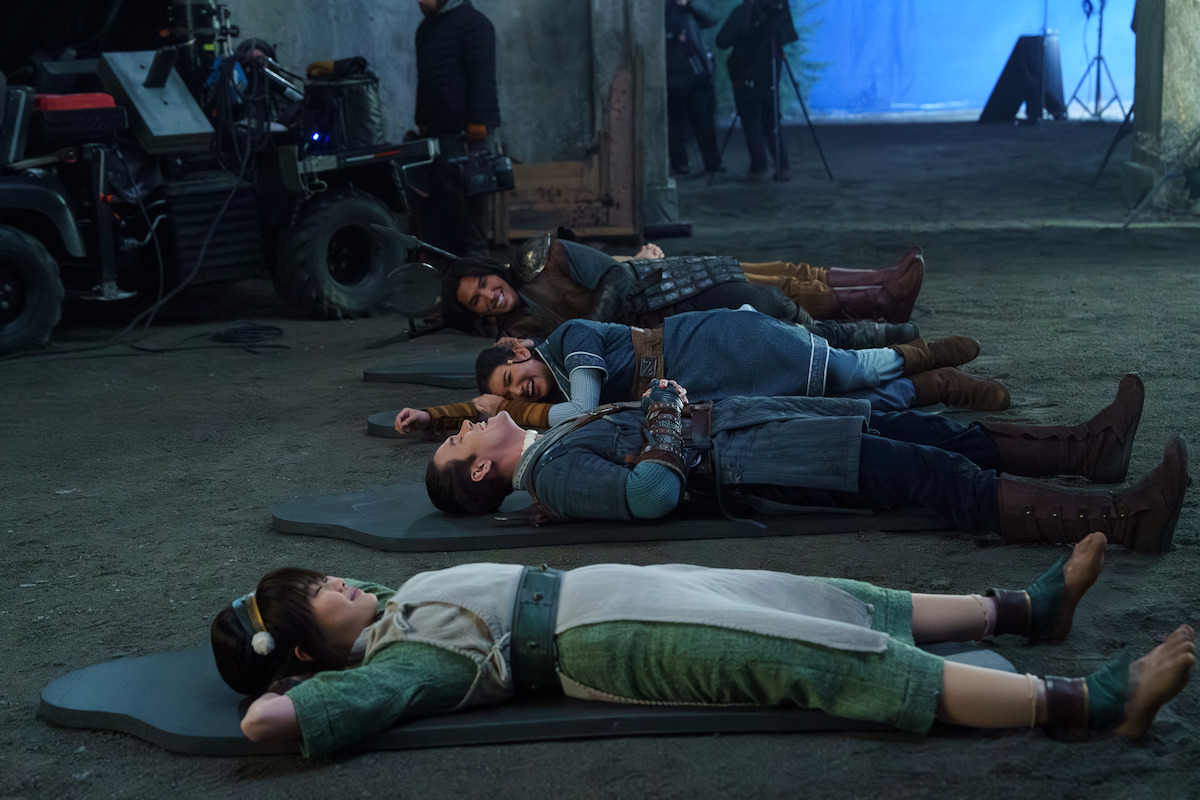 Actors in fantasy costumes lying on mats on a film set with equipment and crew visible, inside an industrial-looking environment, during a break or between scenes. Miyako as Toph wears fake feet shoes.