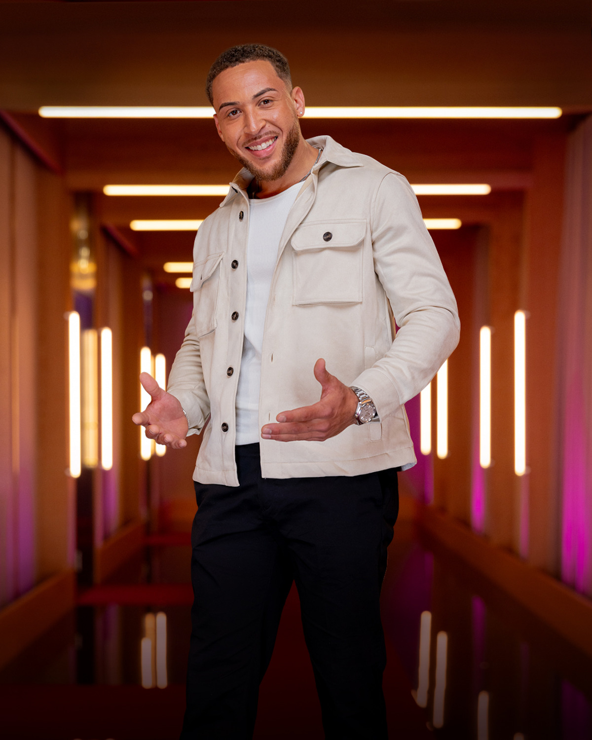 Smiling man standing in a modern, warmly lit hallway with vertical lights on the walls, wearing a light jacket, white shirt, and dark pants, gesturing with his hands.