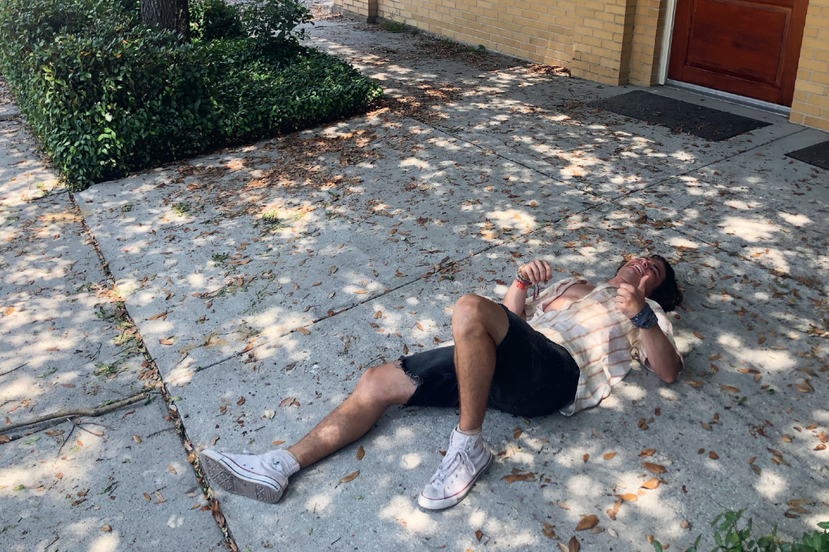 Person lying on a shaded sidewalk under a tree, near a brick building with a wooden door, scattered leaves on the ground, wearing a plaid shirt, shorts, and white sneakers, possibly resting or relaxing outdoors.