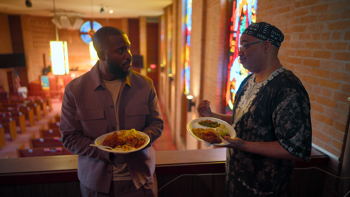 Two men holding plates of food have a conversation in a church, standing by stained glass windows and overlooking empty pews in a warmly lit setting.