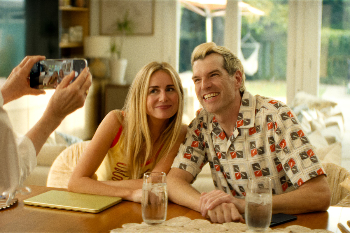 Smiling man and woman sit at a table indoors, posing for a photo being taken by another person. Sunlight streams through large windows, with a bright, cozy home environment in the background. Glasses of water and a laptop are on the table.