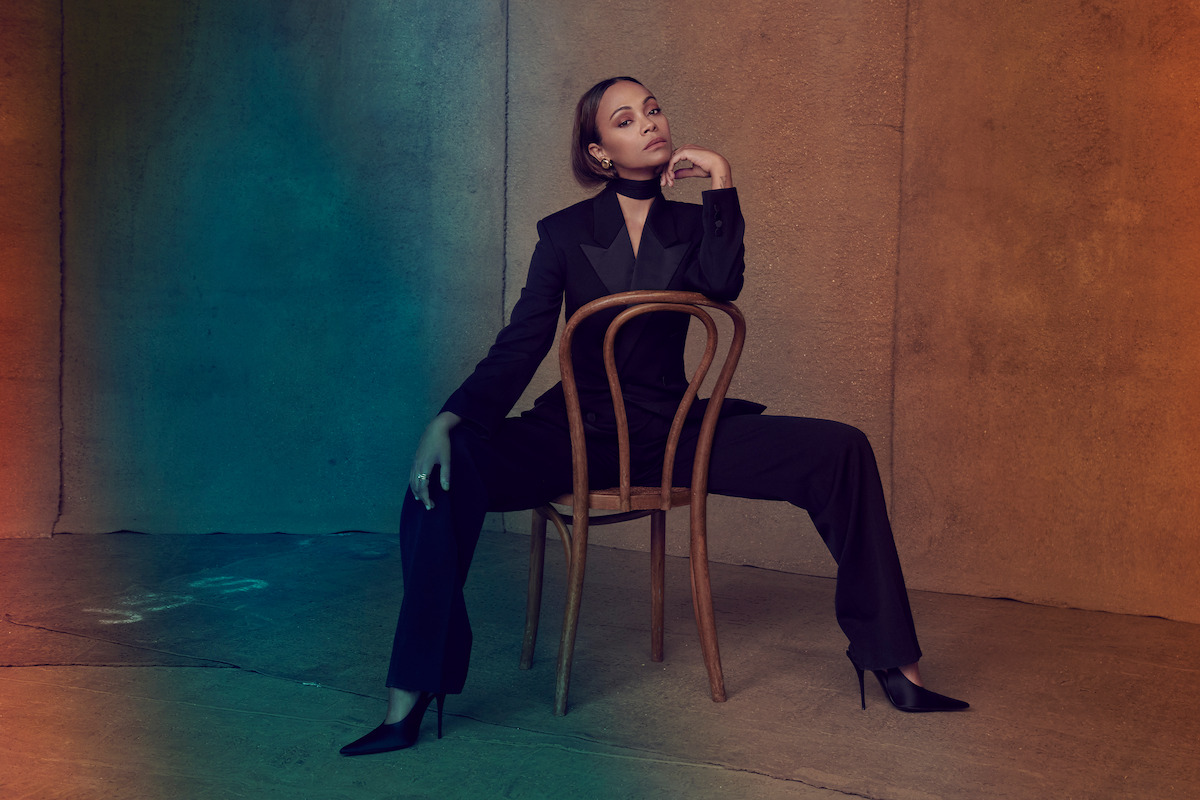 Zoe Saldaña wears a suit and sits backwards in a wooden chair.