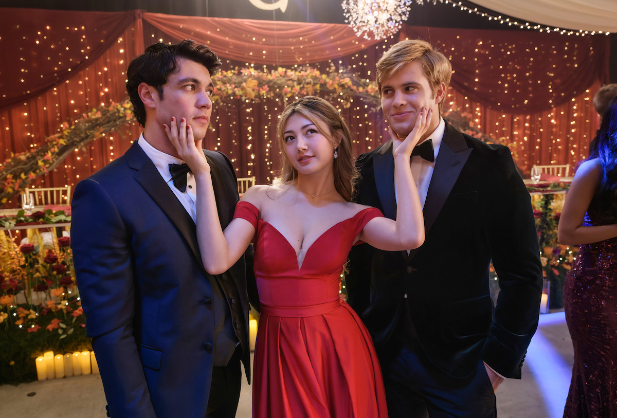 Three young adults dressed formally at an elegant indoor event with string lights and floral decorations, posing for a playful photo; the woman in a red gown stands between two men in suits, touching both of their faces.