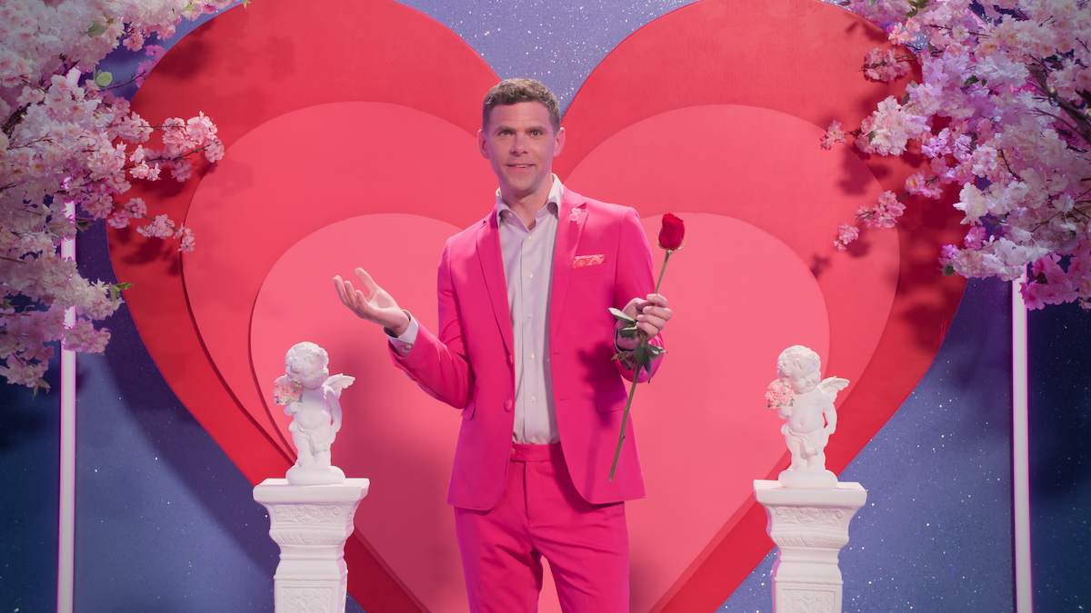 Man in a bright pink suit holding a red rose, standing in front of layered red hearts, surrounded by white cherub statues and pink cherry blossoms on a romantic, vibrant set.