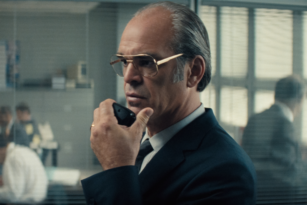 A man in a suit and glasses is holding a walkie-talkie in an office setting with glass walls. Staff are working at desks in the background, and natural light fills the room through large windows.