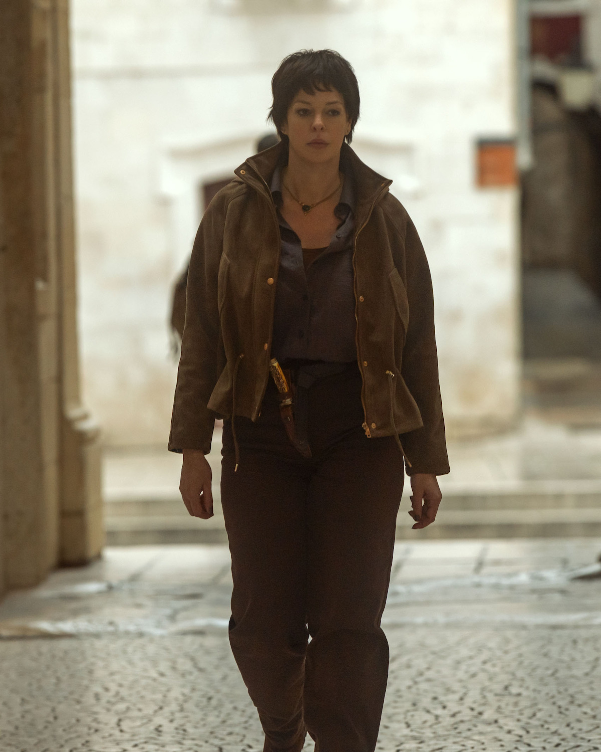 Pollyanna McIntosh as Dalia, with short hair walking alone on a cobblestone street, wearing a brown jacket and dark pants, in an urban outdoor setting with light stone walls and archways in the background.