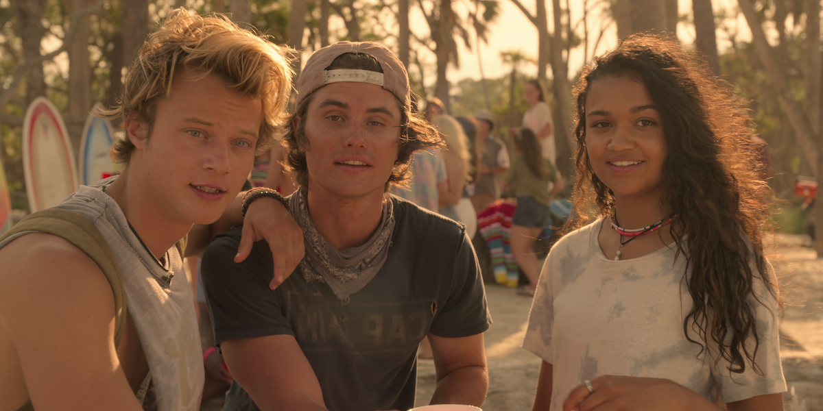 Three teenagers are sitting together outdoors at a beachside gathering, with surfboards and people in the background. The warm lighting suggests sunset, creating a relaxed, summery vibe.