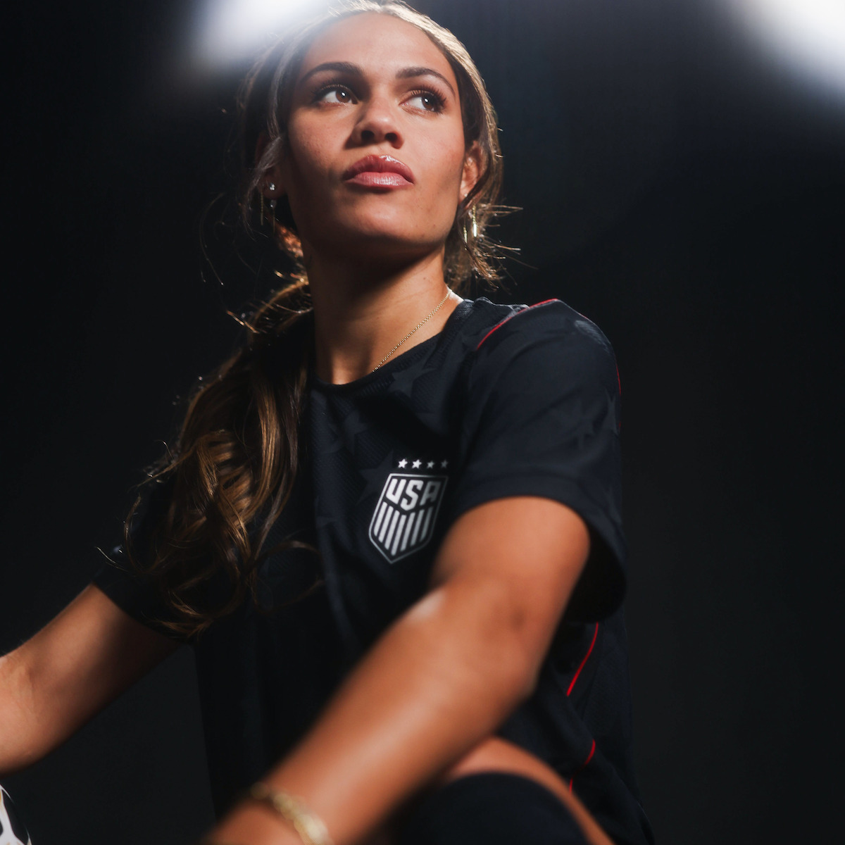 Female soccer player in a black USA jersey sits in a dramatic, low-lit studio setting, looking upward with a confident and focused expression.