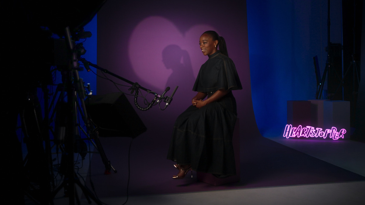 Woman in a black dress sits on a stool in a studio with purple lighting, heart-shaped shadow on the wall, filming equipment visible, and a glowing pink "HEARTSTOPPER" sign in the background.