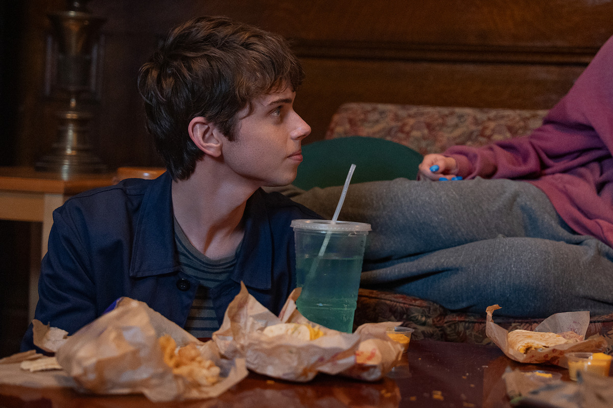 Teenager sitting at a table covered in fast food wrappers and a blue drink, looking to the side in a cozy, dimly lit room with a patterned couch and wood-paneled walls; another person’s legs rest on the couch.