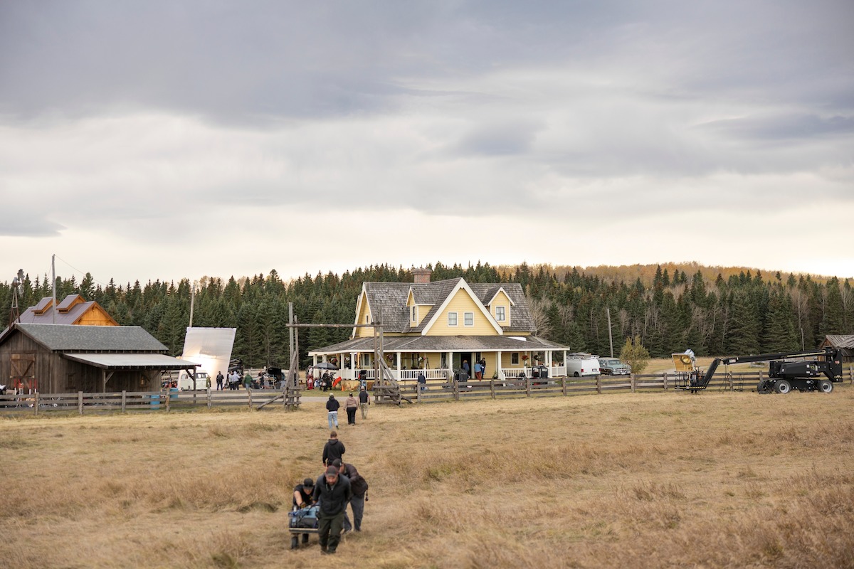 Film crew working in an open field in front of a large yellow farmhouse, surrounded by wooden fences, outbuildings, equipment, and forest trees under a cloudy sky.
