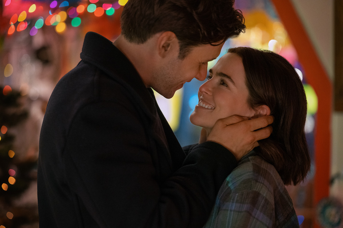 A couple smiling at each other in a loving embrace, standing close together indoors, with festive colorful string lights in the blurry background, creating a warm and romantic atmosphere.