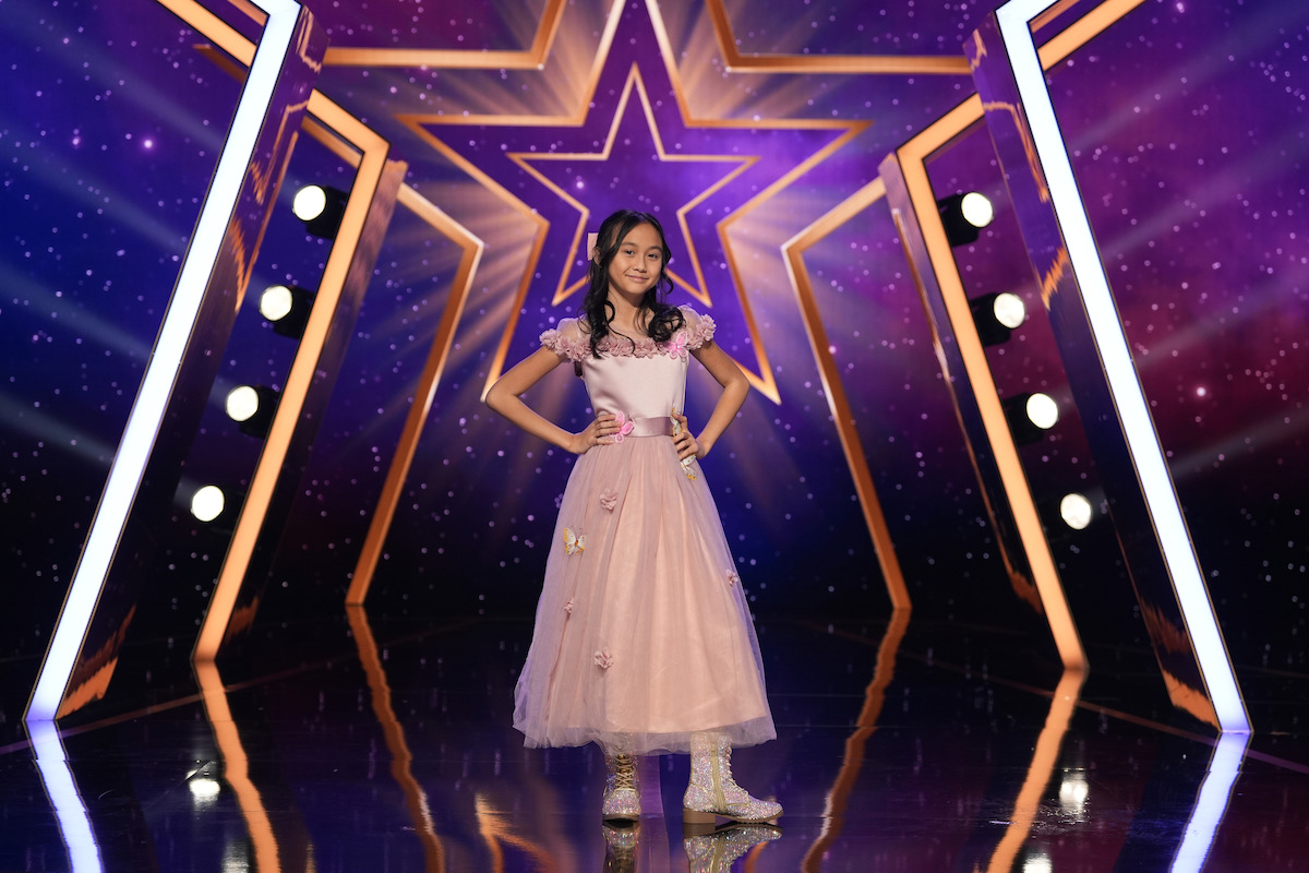 Madison Lauren in a pink dress stands confidently on a shiny stage with a large illuminated star and vibrant purple lighting, suggesting a talent show or performance setting.