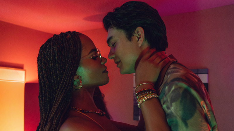 Two people embrace closely in a warmly lit, intimate indoor setting, gazing into each other's eyes. The environment features colorful lighting and a relaxed atmosphere, suggesting a romantic or affectionate moment at a social event.