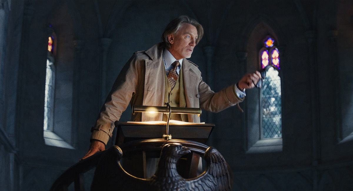 Un hombre mayor vestido con traje formal se encuentra frente a un atril con forma de águila en una habitación de estilo gótico con poca iluminación y vidrieras que crean una atmósfera seria y dramática.