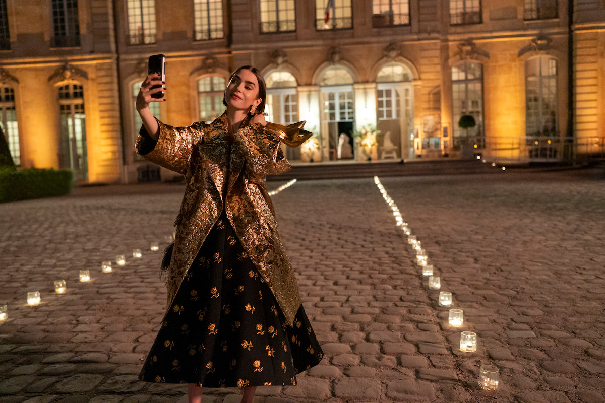 Woman in a gold coat and black dress takes a selfie with her phone at night in a candlelit courtyard outside an elegant, well-lit building with large windows and cobblestone ground.