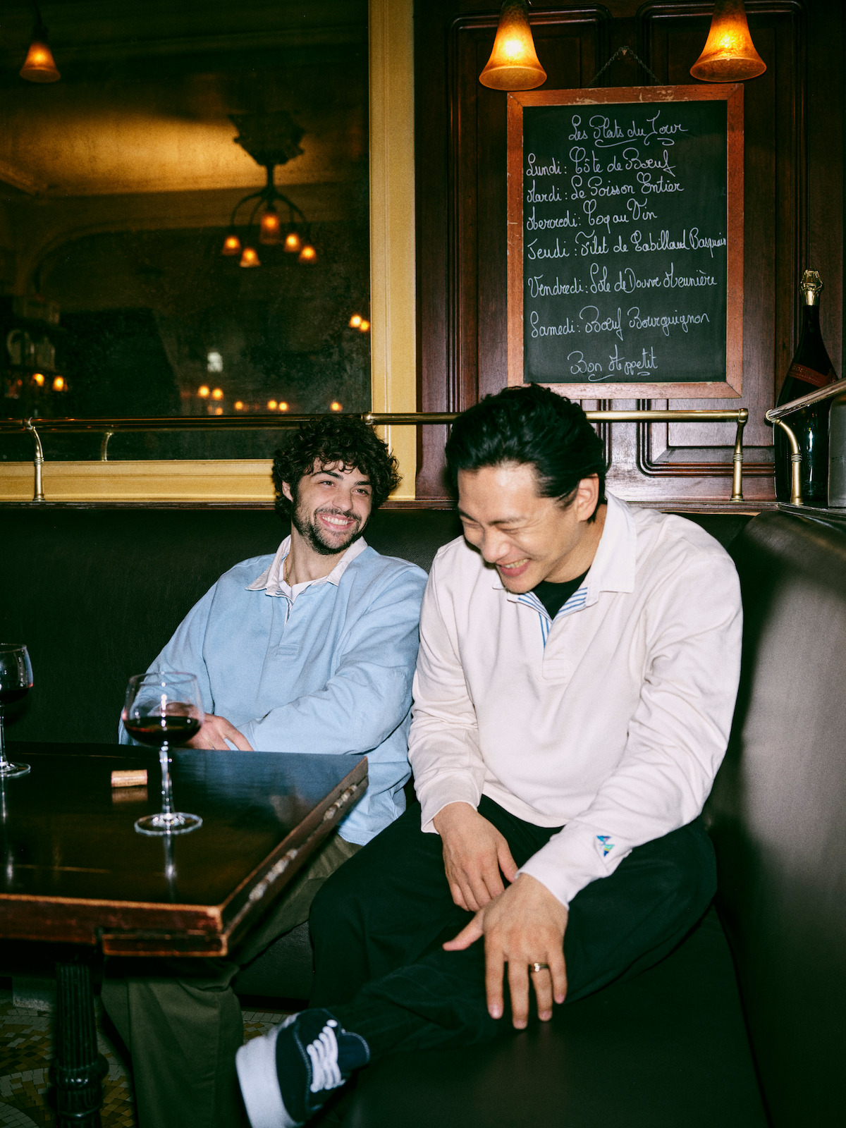 Noah Centineo and Teo Yoo laughing sitting in a restaurant booth over a glass of wine.