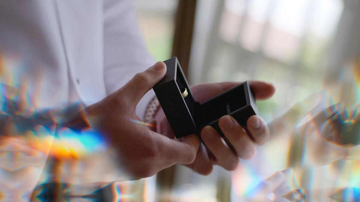A person in a white shirt holds an open black ring box with a ring inside, standing indoors near a window with soft light and colorful light flares in the foreground.