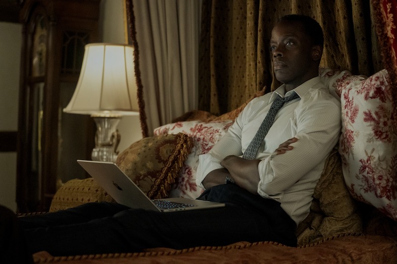 A man in a dress shirt and tie sits on a bed with his arms crossed, looking thoughtful, with a laptop open on his lap in a softly lit, ornate room.