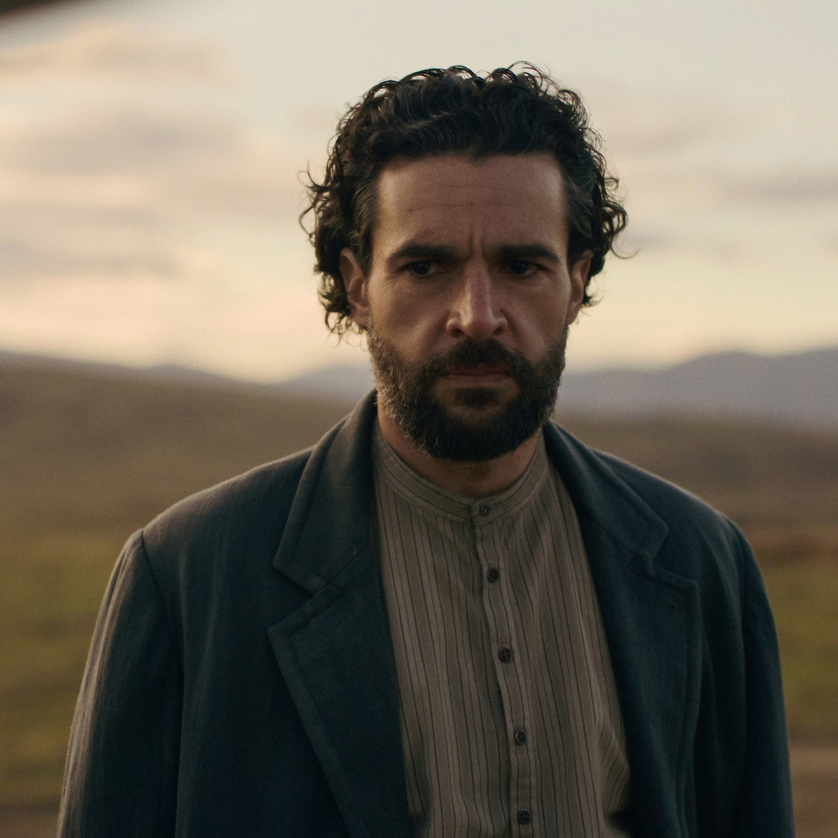 Christopher Abbott como Adam Trask, de cabelos escuros e cacheados, com camisa e casaco de estilo antigo, em pé ao ar livre em uma paisagem rural gramada, com montanhas e a luz suave do entardecer ao fundo.