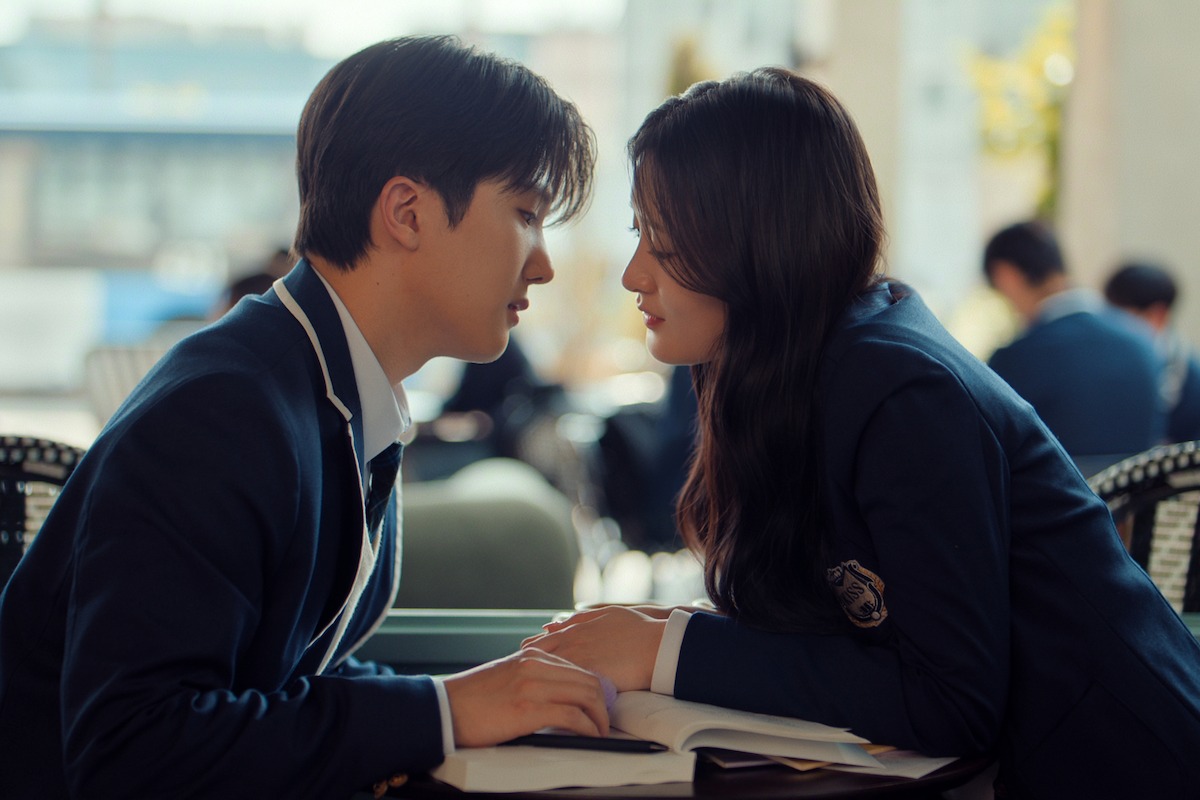 Two students in school uniforms sitting closely at a desk in a bright classroom, leaning toward each other with an open book between them, engaging in a serious and intimate conversation.