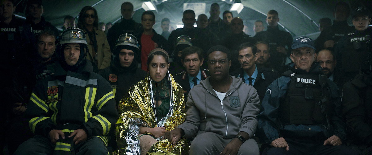 A diverse group of people, including police officers, firefighters, and civilians, sit closely together in what appears to be a dimly lit emergency shelter or tunnel, facing forward with serious and contemplative expressions.