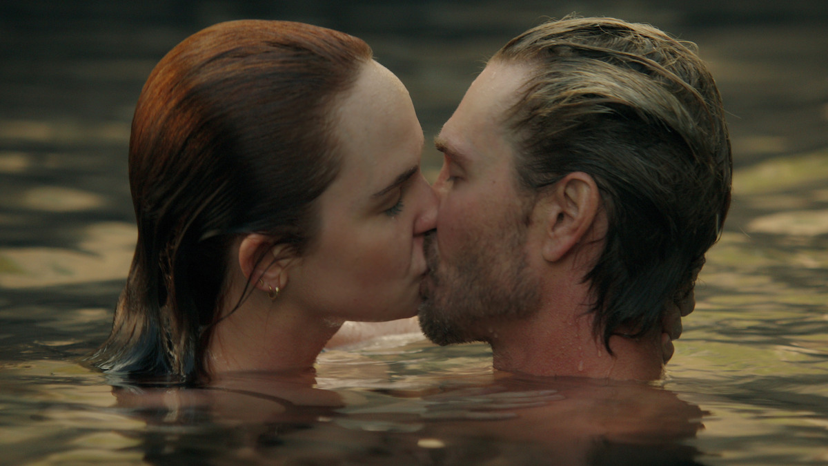 A man and a woman kissing in a body of water, both are partially submerged, with natural lighting and a serene outdoor environment.