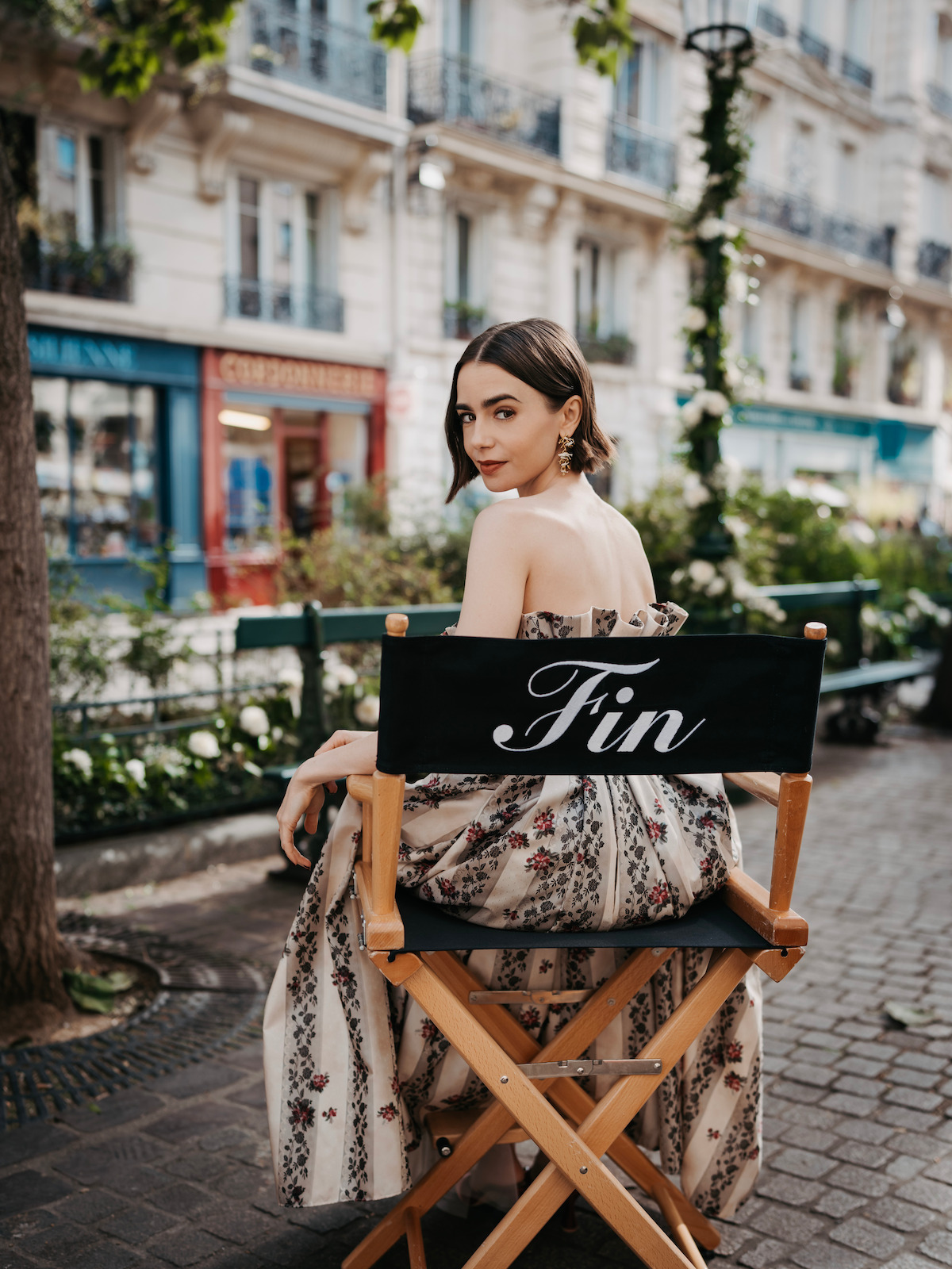 Woman in a floral dress sitting on a director’s chair labeled "Fin" on a Paris street, with quaint buildings, flowers, and shops in the background.