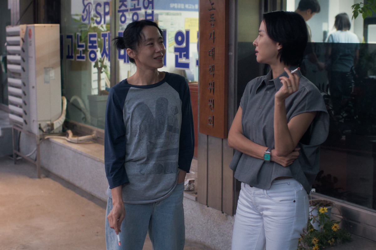 Two women stand outside a building, talking and holding cigarettes. The setting appears urban, with signs in Korean on the windows and a potted plant nearby, suggesting an informal conversation in a city environment.