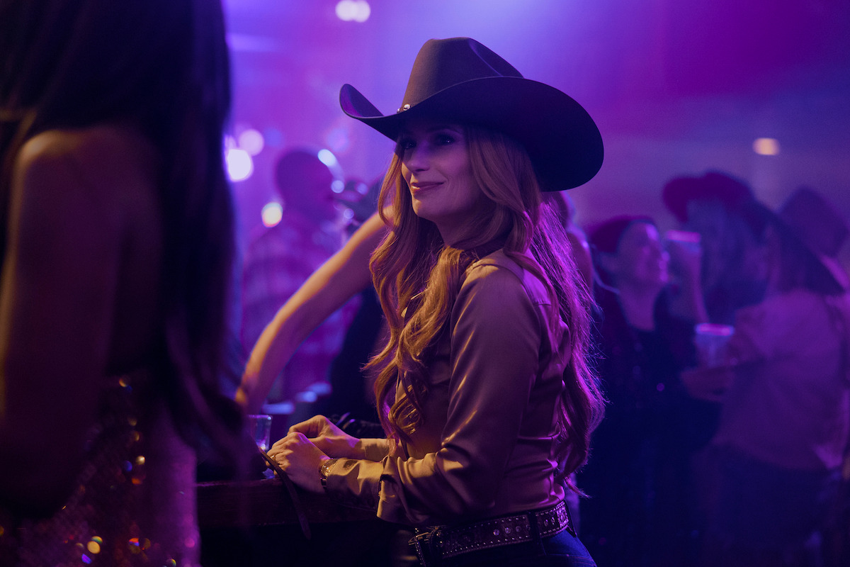 Woman in a cowboy hat and western shirt stands by a bar in a dimly lit, lively nightclub or country bar with people in the background, colorful lights, and a festive atmosphere.