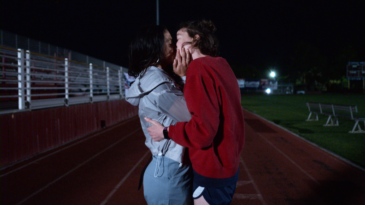 Two people share an intimate kiss on a dimly lit outdoor track at night, with empty bleachers and benches in the background, creating a private and emotional moment.