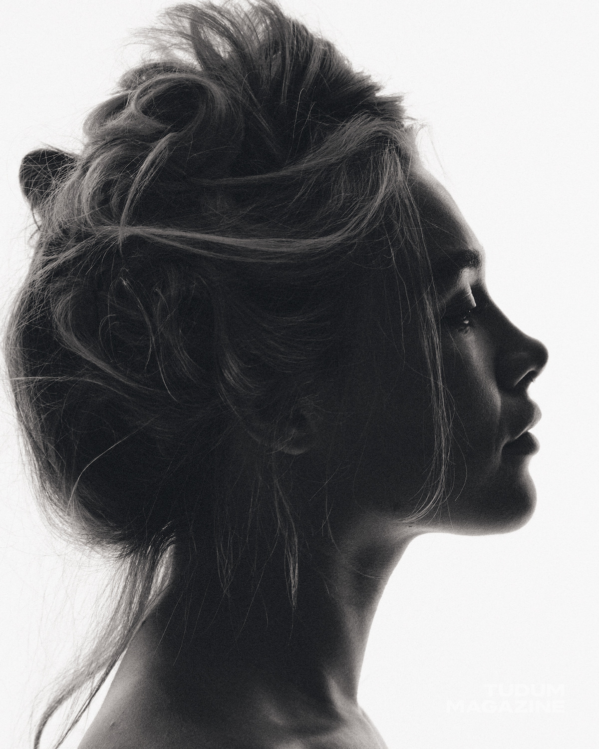 Black and white portrait of a woman in profile with messy hair, styled up. She is against a plain white background, creating a high-contrast, artistic and softly lit setting.