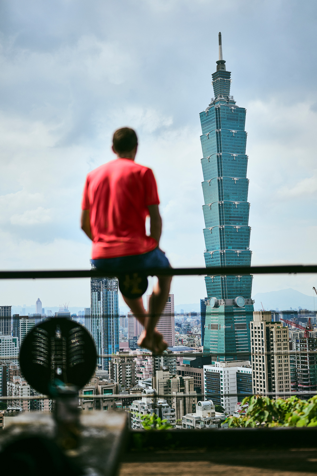 A person in a red shirt sits on a railing overlooking a cityscape with the Taipei 101 skyscraper, under a cloudy sky, creating a peaceful and contemplative mood.