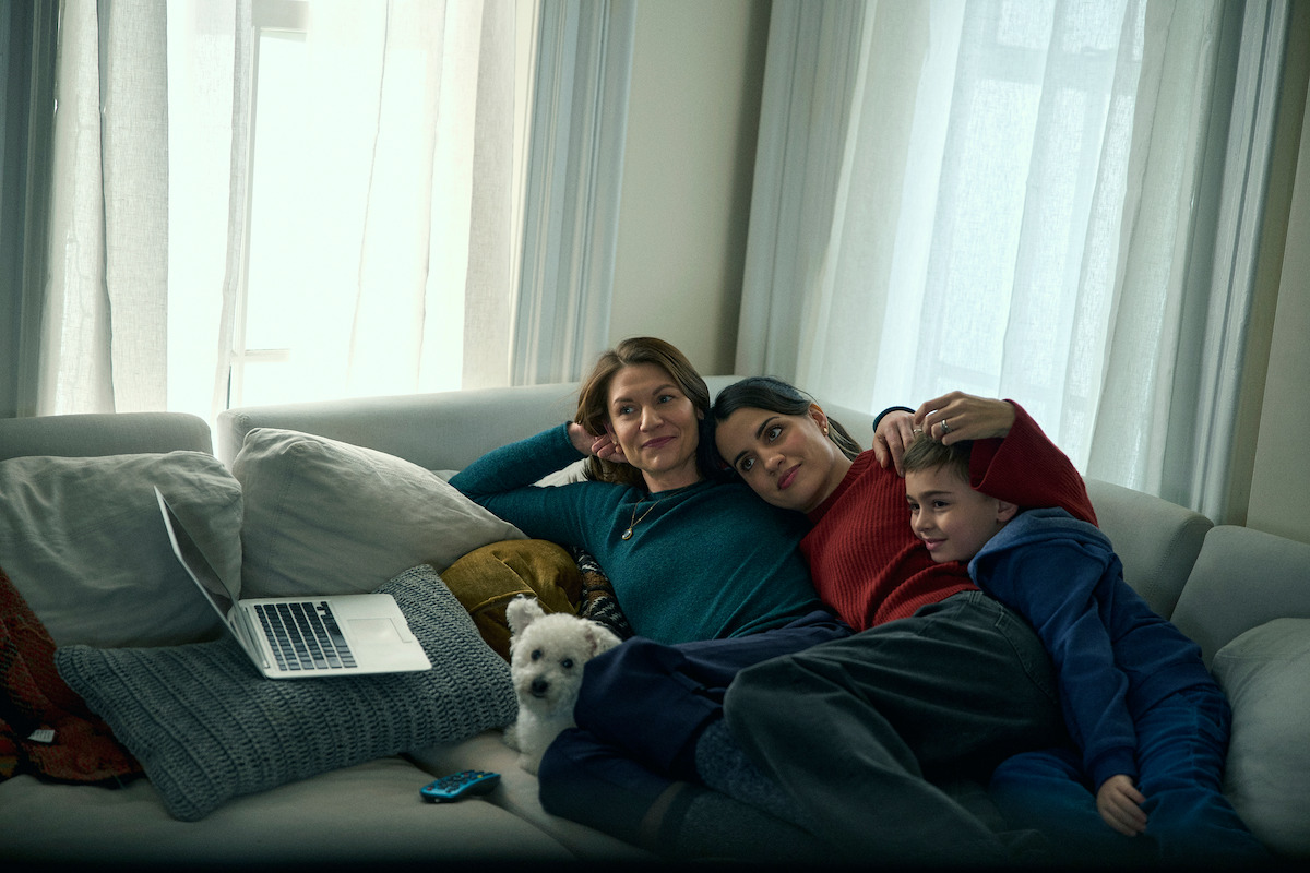 Three people and a small white dog relax together on a cozy couch in a warmly lit living room with a laptop nearby, creating a peaceful, intimate, and comfortable mood.