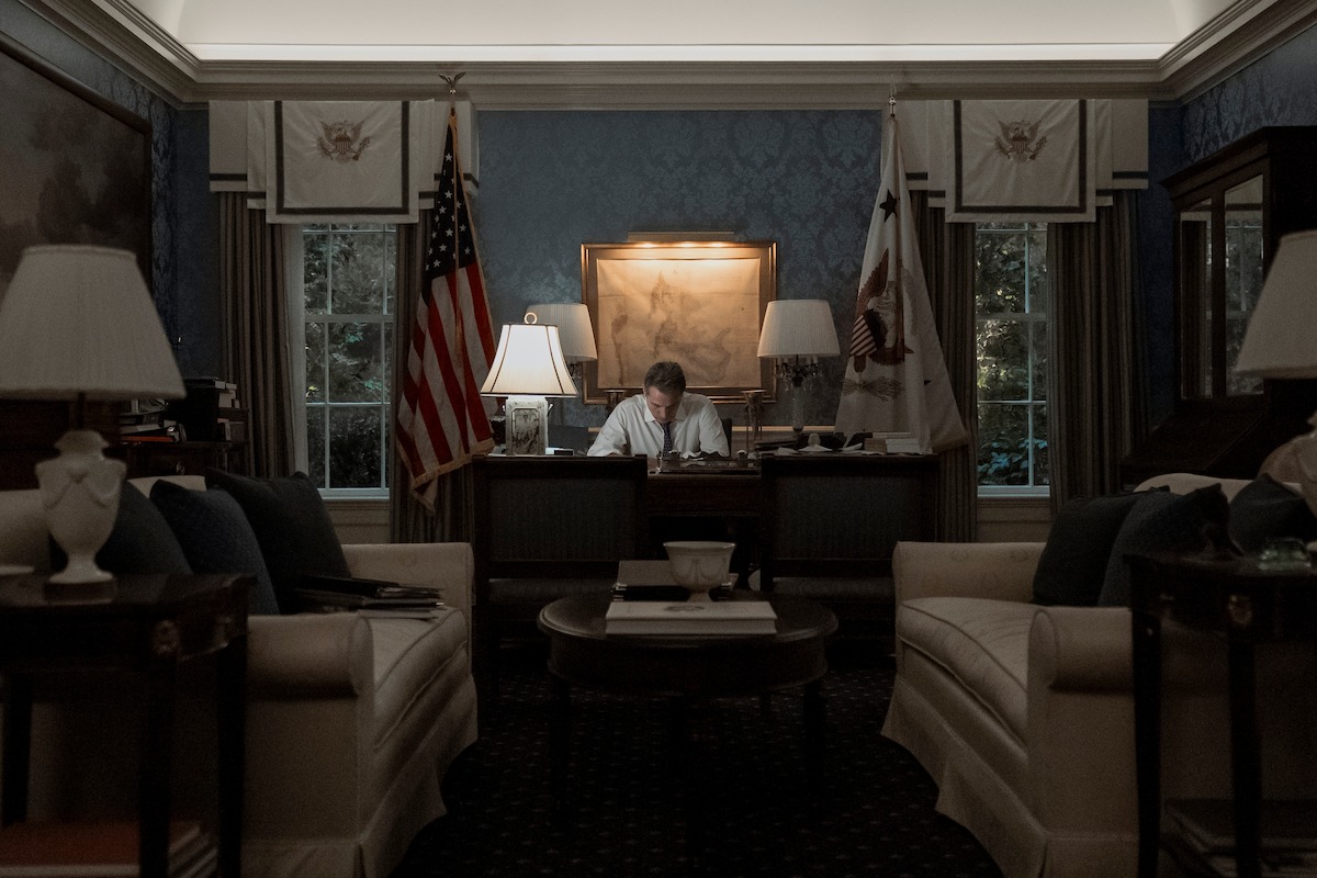 A man works alone at a large desk in a dimly lit, formal office with flags, artwork, and elegant furnishings, creating a serious, contemplative mood.