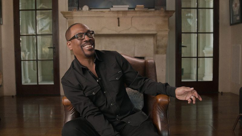 Eddie Murphy wears a black jacket and smiles looking away from the camera.