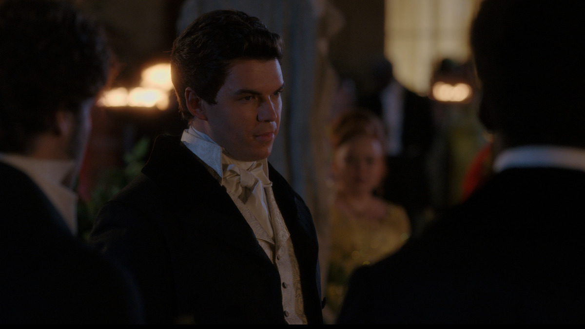 A man in formal 19th-century attire stands in a dimly lit ballroom, surrounded by other guests dressed elegantly, with warm lights and soft background details enhancing the period setting.