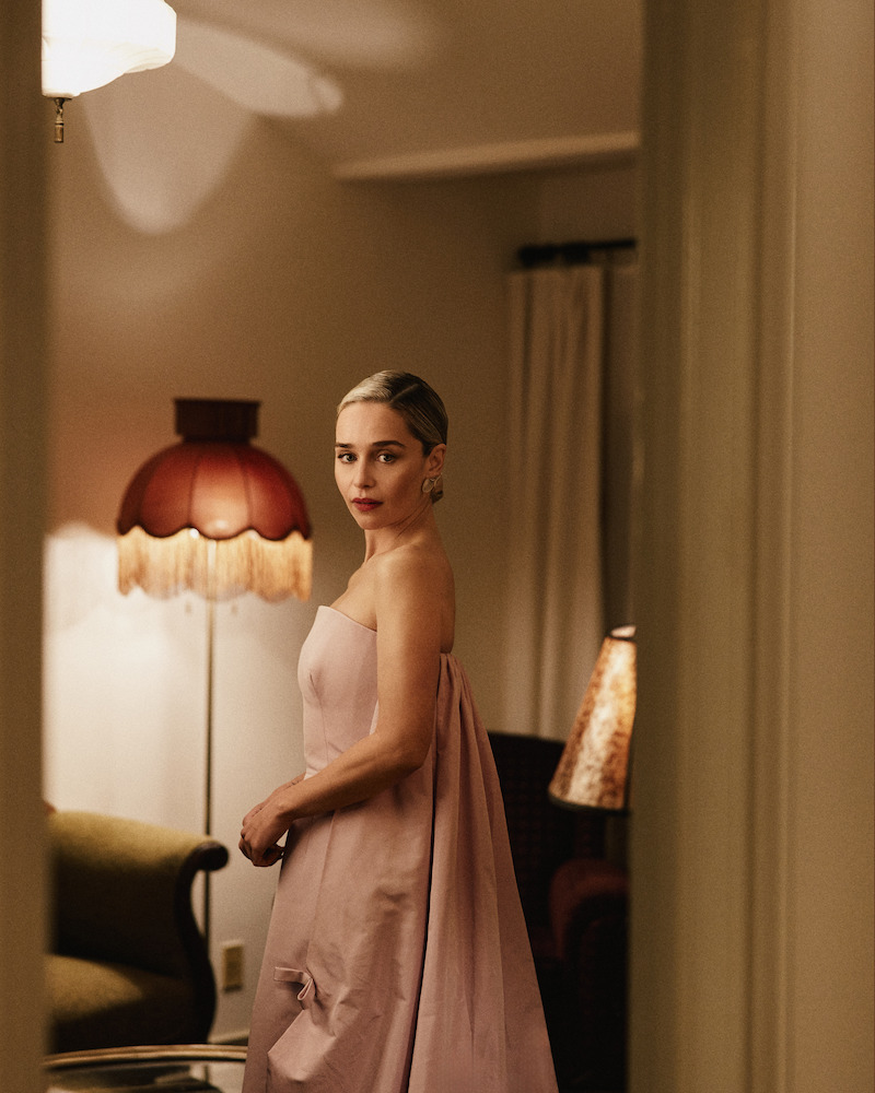Woman in an elegant pink dress stands in a warmly lit vintage-style living room with retro lamps, beige walls, and classic furniture, looking thoughtfully towards the camera.