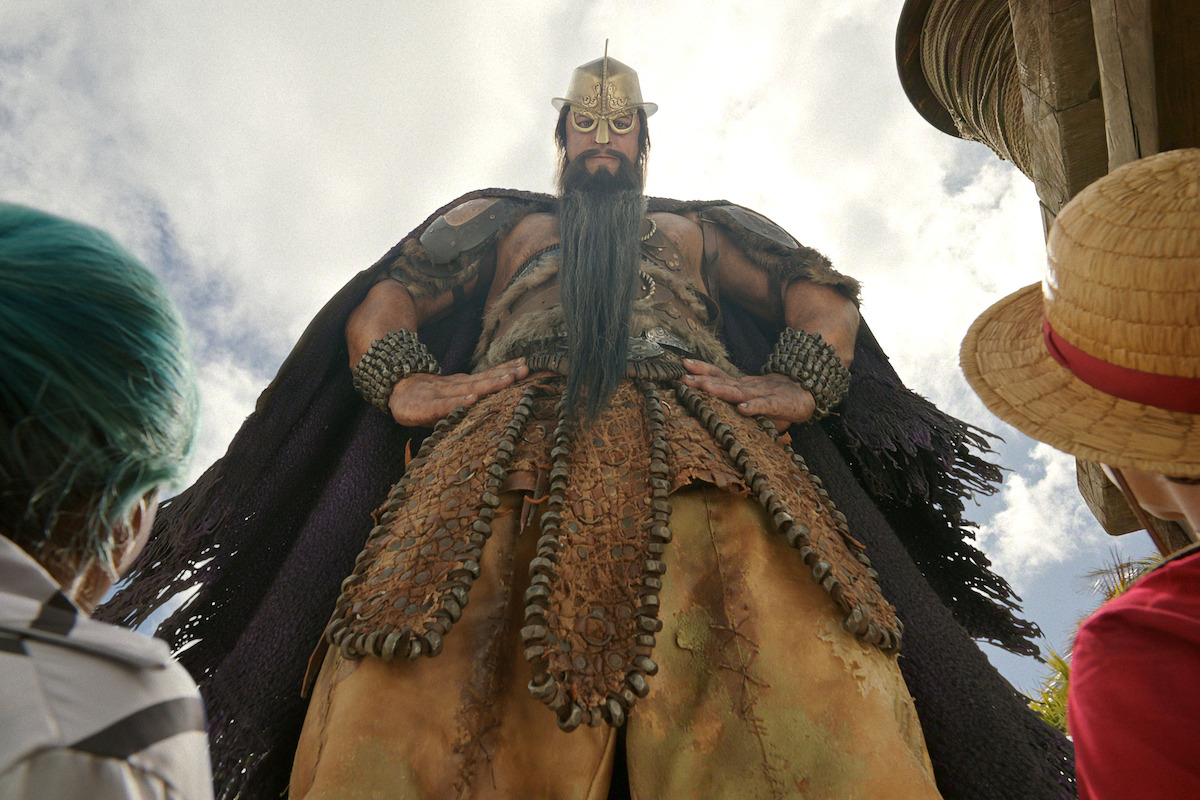 A towering armored warrior with a helmet and long beard stands confidently outdoors, looking down at two people with colorful hair and hats under a cloudy sky, giving a dramatic and imposing scene.