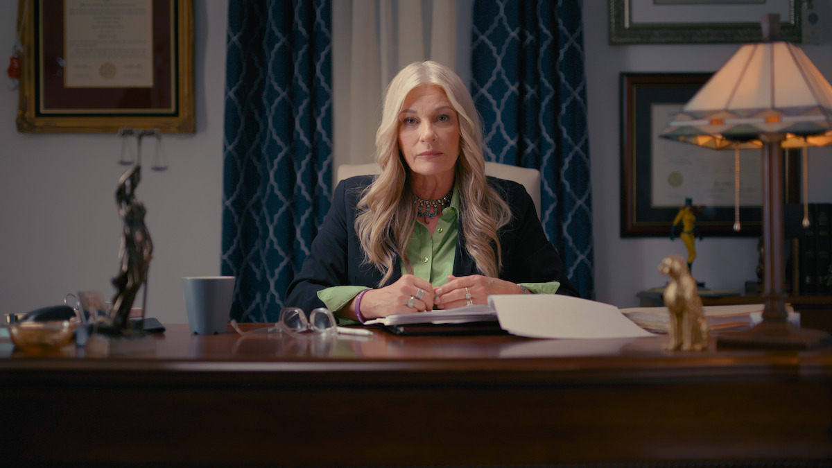 A woman with long blonde hair sits at an office desk with papers, a lamp, and legal decorations, in front of blue curtains and framed certificates, appearing professional and attentive.