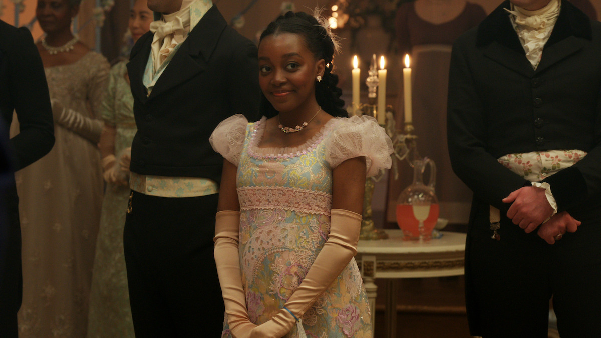 Young woman in a pastel Regency-era gown smiles at a formal ball, surrounded by elegantly dressed guests. Candles and a table with a jug of punch create a warm, historical atmosphere.