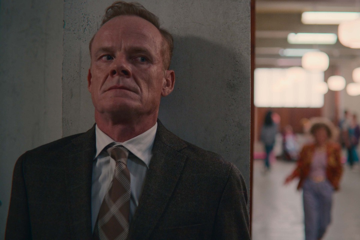 Alistair Petrie as Michael Groff hides behind a wall in a hallway in Season 4 of ‘Sex Education’