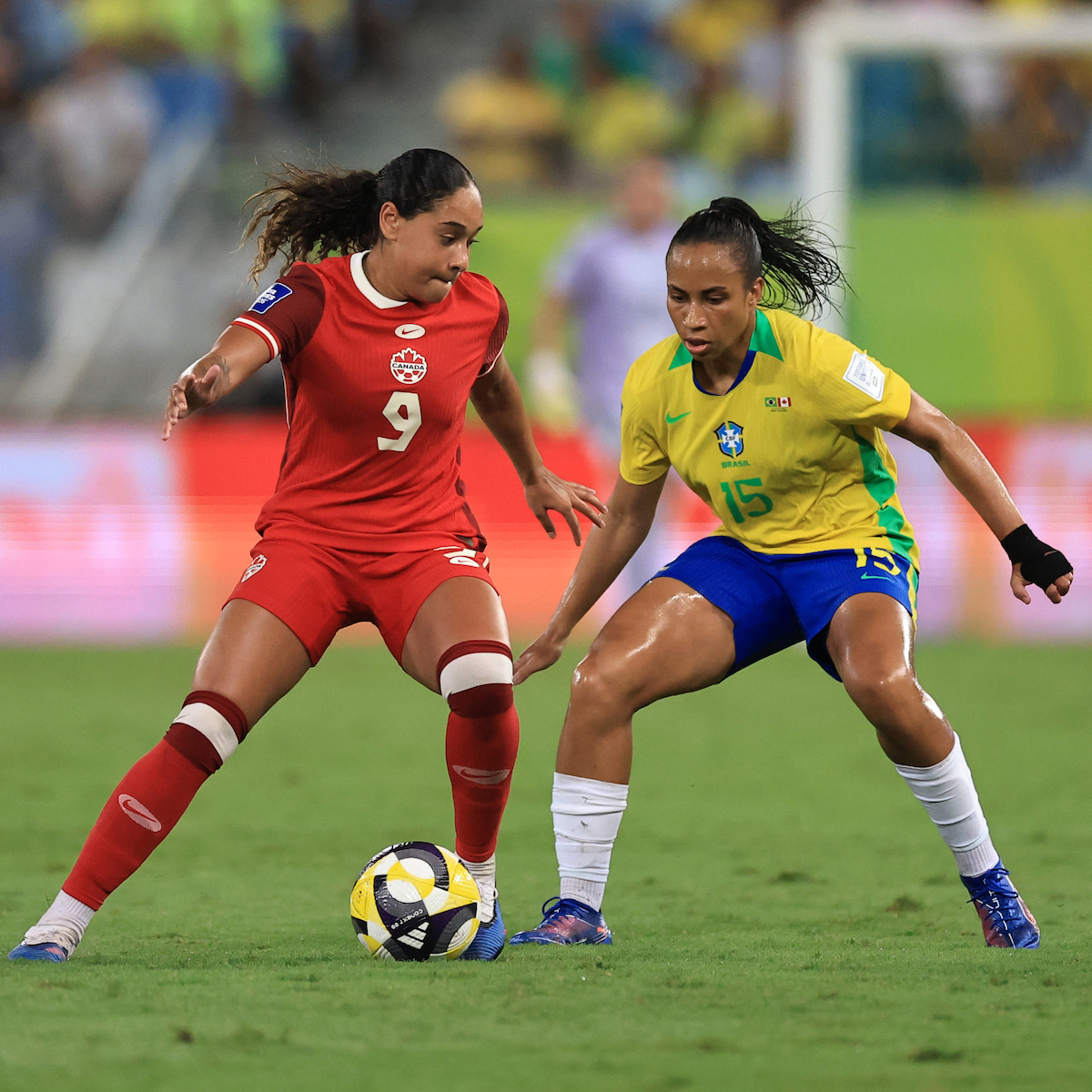 Two female soccer players compete for possession of the ball on a grass field during a match, with one wearing a red Canada kit and the other in a yellow and blue Brazil kit. Stadium and spectators blurred in the background.